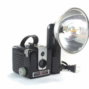 May include: A black Brownie Hawkeye camera with a flash attachment. The flash has a silver reflector and a clear light bulb. The camera has a black leather strap and a silver knob on the side.