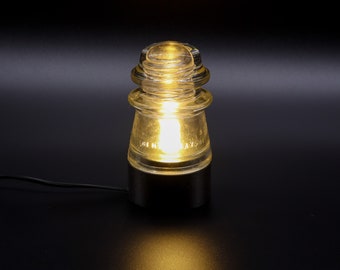 May include: A glass lamp with a black base, made from an old insulator. The lamp is lit and casting a warm glow.