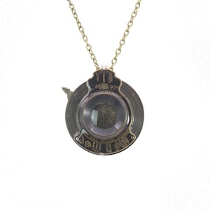 May include: A vintage-style pendant necklace featuring a circular, dark metal pendant with a central lens. The pendant has engraved text and is suspended from a delicate gold-colored chain. The design evokes a classic, industrial aesthetic.