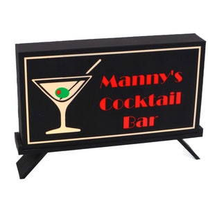 May include: Black sign with a white border and a martini glass graphic. The sign reads "Manny's Cocktail Bar" in red letters.