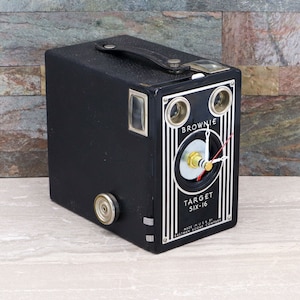 Puede incluir: Una cámara vintage negra Brownie Target Six-16 convertida en reloj. La cámara tiene una cara plateada con las palabras "Brownie" y "Target Six-16" impresas en ella. La esfera del reloj es negra con manecillas blancas y una perilla plateada en el centro.
