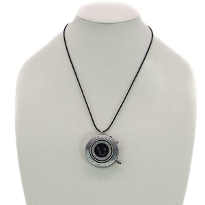 Puede incluir: Un collar con un colgante de lente de cámara plateado con un cordón negro. La lente tiene un diseño negro y plateado con el texto "Kodak Flash 200 Shutter" impreso en el lateral.