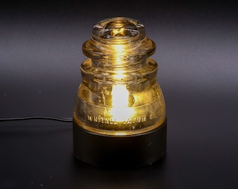 May include: A vintage glass insulator lamp with a black base. The insulator is amber colored and has the text "WHITTALL LATUM CO" on the side.