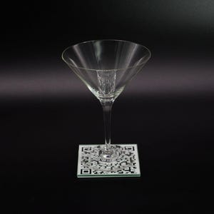 May include: A clear glass martini glass sits on a square coaster with a black and white QR code design.