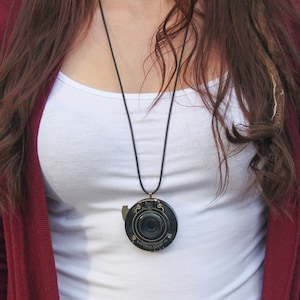 May include: A woman wearing a white tank top and a maroon cardigan is wearing a necklace with a vintage camera lens pendant. The lens is black with gold accents and has the text "PATENTED SEPT 21 1914" on it.