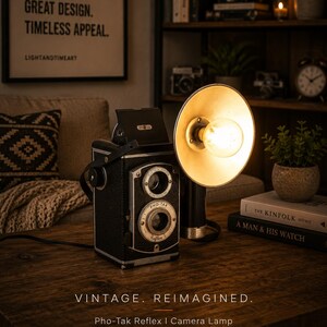 May include: A vintage Pho-Tak Reflex I camera repurposed into a functional table lamp. The black camera body features a silver lens housing and is attached to a circular, metallic reflector shade containing a glowing, warm-toned light bulb.