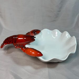 May include: A white ceramic bowl with a scalloped edge, featuring a glossy red lobster figurine attached to the side. The lobster is detailed with dark red shading and extends outward from the rim of the bowl, creating a decorative accent.