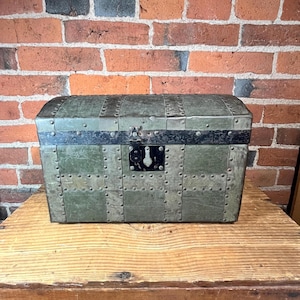 May include: A rectangular storage trunk with a domed lid, featuring a muted olive green finish. The exterior is reinforced with dark metal bands and numerous rivets, and it includes a central black metal latch mechanism with a keyhole plate.