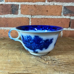 May include: A white ceramic teacup featuring a deep blue transferware pattern depicting a landscape with trees, buildings, and a bridge. The interior rim is decorated with a solid blue band, and the handle has a circular loop design.