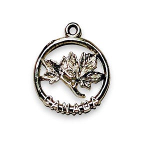 May include: A silver-colored pendant featuring a detailed maple leaf design within a circular frame. The word "CANADA" is inscribed along the bottom edge of the circle. The pendant has a small loop at the top for attaching to a chain.
