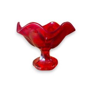May include: A vibrant red glass bowl featuring a ruffled, scalloped rim and a pedestal base. The translucent material catches the light, highlighting the deep crimson hue and the smooth, curved edges of the decorative footed dish.