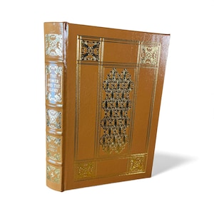 May include: A hardcover book with a brown leather cover and gold embossed decorative elements. The spine features the title "THE POWER AND THE GLORY" and the author's name. The book's cover has a central ornate design and gold border details.