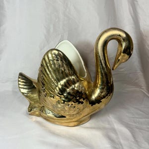 May include: A decorative swan-shaped planter or vessel featuring a highly reflective metallic gold finish on the exterior and a smooth, solid white interior. The swan is depicted with its neck curved downward and wings slightly raised.
