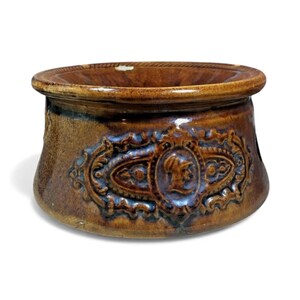 May include: A brown ceramic bowl with a decorative embossed design. The bowl features a detailed relief of a person's profile within an ornate frame. The rim has a rope-like texture. The bowl has a glossy finish.