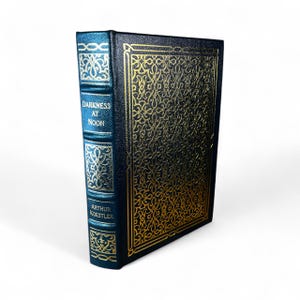 May include: A hardcover book titled "Darkness at Noon" by Arthur Koestler. The book has a dark blue cover with gold filigree and gold lettering. The spine is dark blue with gold accents and the title and author's name are visible.