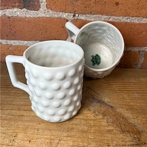 May include: Two white ceramic mugs with a textured, dotted surface. One mug is upright, while the other is tilted, revealing a green frog design at the bottom. Both mugs have square handles and are set against a brick wall and wooden surface.
