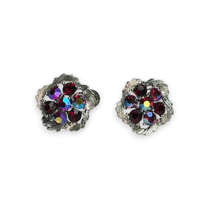 May include: A pair of vintage flower-shaped stud earrings. Each earring features a cluster of deep red and iridescent crystals set in a silver-tone metal frame with a textured, leaf-like design. The earrings are displayed on a white background.
