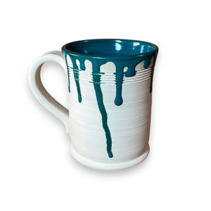 May include: White ceramic mug with a teal interior and teal paint drips. The mug has a large handle and a textured exterior. The paint drips down the side of the mug, creating a unique design. This handmade mug is perfect for coffee or tea.