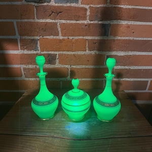 May include: Two tall glass decanters with matching stoppers and one round glass jar with a lid, all glowing a bright neon green color. Each piece features a decorative gold-colored band with a repeating pattern around the center section.