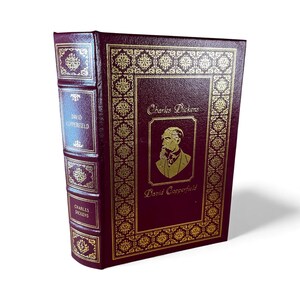May include: A maroon hardcover book titled "David Copperfield" by Charles Dickens. The cover features gold embossed decorative patterns and a portrait of the author. The spine also has the title and author's name in gold.