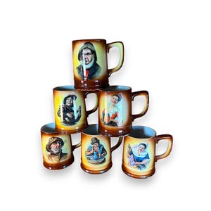 May include: A stack of six brown ceramic mugs with painted portraits of people. Each mug has a handle and a different portrait, including a fisherman, a woman, and a man in a hat. The mugs are arranged in a pyramid shape.