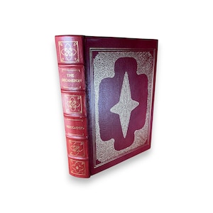 May include: A hardcover book titled "The Decameron" by Boccaccio. The book has a burgundy leather cover with gold embossed details and a gold starburst design. The spine is red with gold lettering and decorative elements.