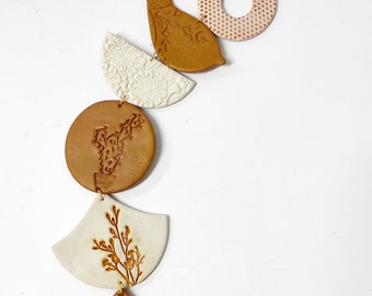 May include: A brown and white ceramic wall hanging with a bird, cactus, and floral designs. The hanging is attached to a brown cord.