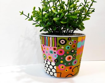 May include: A decorative, hand-painted flower pot with a vibrant patchwork design. The pot features a mix of colors, including pink, green, orange, and blue, with a variety of patterns. The pot is filled with a green plant.