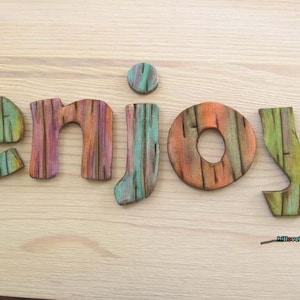 Könnte beinhalten: Holzbuchstaben mit dem Wort "enjoy" in verschiedenen Farben, darunter Grün, Orange, Rosa und Blau. Die Buchstaben haben eine Holzmaserung und sind auf einer hellen Holzoberfläche angeordnet. Das Wort ist ein dekoratives Element.