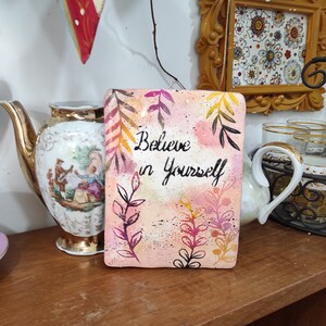 Könnte beinhalten: Ein rosafarbenes und weißes bemaltes Holzschild mit den Worten "Believe in Yourself" in schwarzer Kursivschrift. Das Schild ist mit schwarzen, rosa und gelben floralen Designs verziert.
