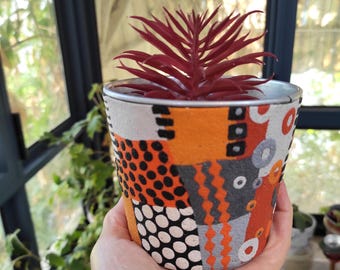 May include: A decorative plant pot with a vibrant, geometric pattern in orange, gray, black, and white. The pot holds a burgundy-colored succulent plant. The pot has a silver rim and is held in a hand.
