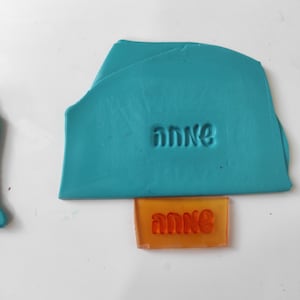 May include: A teal blue polymer clay block with the word "anne" embossed into the surface. Below the block is an orange rubber stamp with the same word, "anne", in a raised font.
