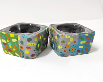 May include: Two square ceramic candle holders with a colorful, abstract design. The holders are gray with green, blue, yellow, orange, and red accents.