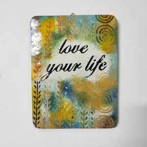 May include: A rectangular wooden sign with a white background and a green, blue, and yellow abstract design. The sign has the words "love your life" written in black paint.