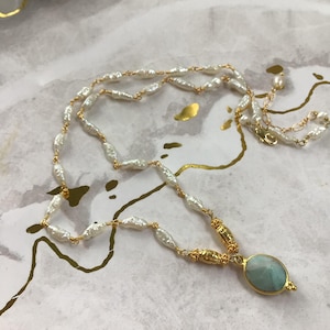 May include: A delicate necklace featuring white, oblong pearls strung on gold wire. The necklace includes a gold pendant with a light blue oval stone. The jewelry rests on a white and gold patterned surface.