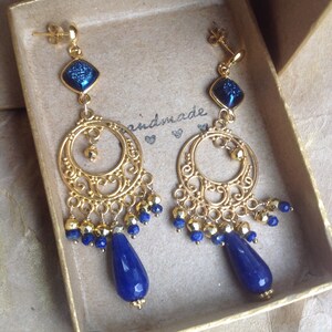 Puede incluir: Un par de pendientes dorados con detalles azules. Cada pendiente presenta una gema azul cuadrada, un diseño de filigrana y gemas azules en forma de lágrima colgantes. Los pendientes se presentan en una caja dorada. La palabra 'handmade' es visible.