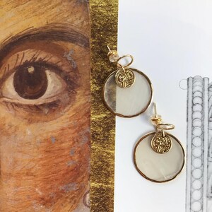 Könnte beinhalten: Goldfarbene Ohrringe mit kreisförmigem Design. Jeder Ohrring hat einen klaren, kreisförmigen Rahmen mit einem weißen, halbtransparenten Mittelteil und einem kleinen Goldmünzen-Anhänger. Die Ohrringe werden vor einem weißen und goldenen Hintergrund gezeigt.
