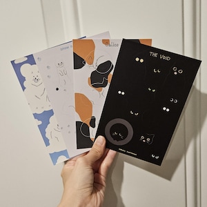 Op de afbeelding: Een hand houdt vier kunstprints met kattenillustraties vast. De prints bevatten een blauw-wit wolkenontwerp met een hond, een witte print met een kat, een lapjeskatontwerp en een zwarte print met de titel "THE VOID" met kattenogen.