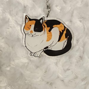 Op de afbeelding: Een sleutelhanger in de vorm van een calico kat met een wit, oranje en zwart patroon. De kat zit met zijn ogen dicht en een glimlach op zijn gezicht. De sleutelhanger is gemaakt van helder acryl en heeft een zilveren metalen ring.