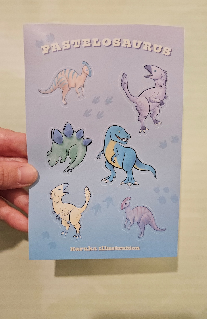 Op de afbeelding: Een lichtblauwe kaart met cartoon-dinosaurussen in verschillende kleuren. Het woord "PASTELOSAURUS" staat bovenaan gedrukt. De naam van de kunstenaar, "Haruka illustration", staat onderaan gedrukt.