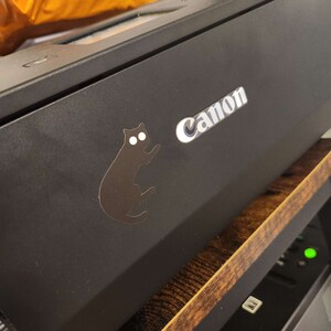 Op de afbeelding: Zwarte Canon-printer met een zilveren logo en een zwarte kattensticker. De printer staat op een houten plank. Op de achtergrond is een oranje pakket zichtbaar.