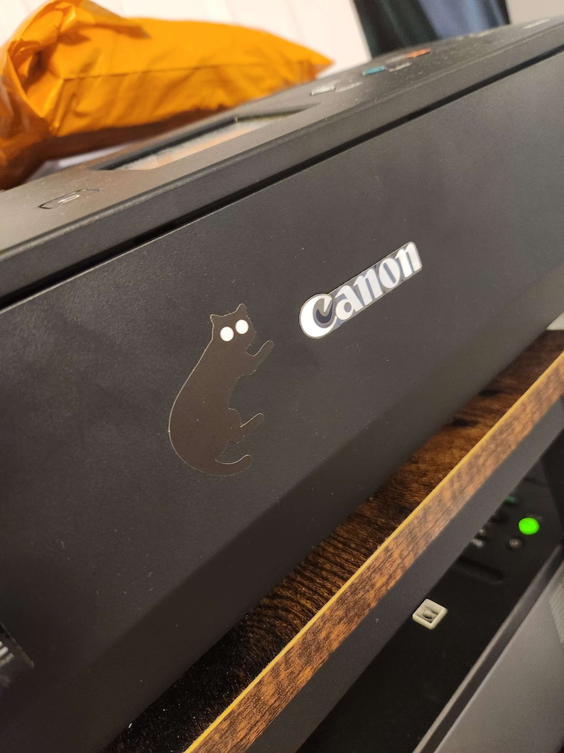 Op de afbeelding: Zwarte Canon-printer met een zilveren logo en een zwarte kattensticker. De printer staat op een houten plank. Op de achtergrond is een oranje pakket zichtbaar.