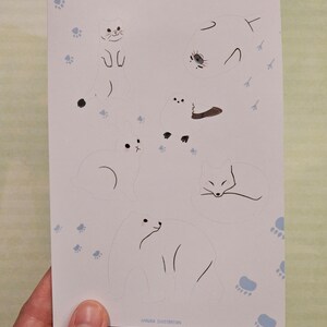 Op de afbeelding: Een witte kaart met illustraties van sneeuwdieren, waaronder een kat, een zeehond, een konijn, een vos en een ijsbeer. De woorden "snow animals" zijn in lichtblauw geschreven. Lichtblauwe pootafdrukken en de woorden "HANUKA ILLUSTRATION" zijn ook zichtbaar.