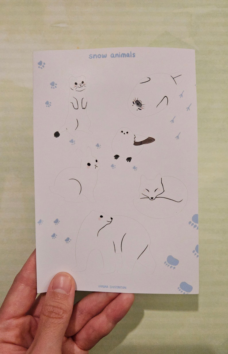 Op de afbeelding: Een witte kaart met illustraties van sneeuwdieren, waaronder een kat, een zeehond, een konijn, een vos en een ijsbeer. De woorden "snow animals" zijn in lichtblauw geschreven. Lichtblauwe pootafdrukken en de woorden "HANUKA ILLUSTRATION" zijn ook zichtbaar.