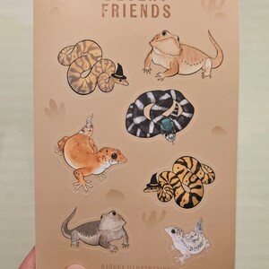 Op de afbeelding: Een bruin kaartje met illustraties van woestijnreptielen, waaronder slangen en hagedissen, sommige met hoeden. De tekst "DESERT FRIENDS" staat bovenaan. De naam van de kunstenaar, "HARUKA ILLUSTRATION", staat onderaan.