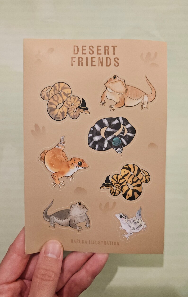 Op de afbeelding: Een bruin kaartje met illustraties van woestijnreptielen, waaronder slangen en hagedissen, sommige met hoeden. De tekst "DESERT FRIENDS" staat bovenaan. De naam van de kunstenaar, "HARUKA ILLUSTRATION", staat onderaan.
