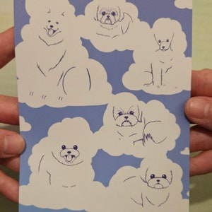 Op de afbeelding: Een blauwe ansichtkaart met illustraties van honden in de vorm van pluizige wolken. De tekst "Fluffy Clouds" staat bovenaan, met "HARUKA ILLUSTRATION" onderaan. De honden zijn getekend in een eenvoudige lijnstijl.