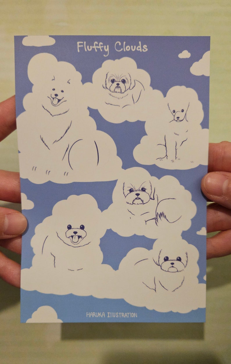 Op de afbeelding: Een blauwe ansichtkaart met illustraties van honden in de vorm van pluizige wolken. De tekst "Fluffy Clouds" staat bovenaan, met "HARUKA ILLUSTRATION" onderaan. De honden zijn getekend in een eenvoudige lijnstijl.