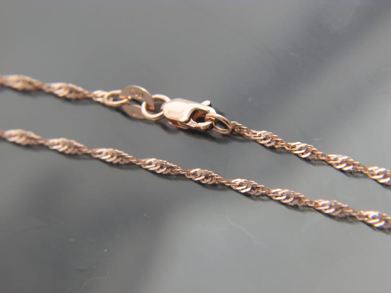 May include: A rose gold necklace with a delicate twisted rope chain design. The chain is secured with a lobster clasp. The necklace is displayed against a dark background.