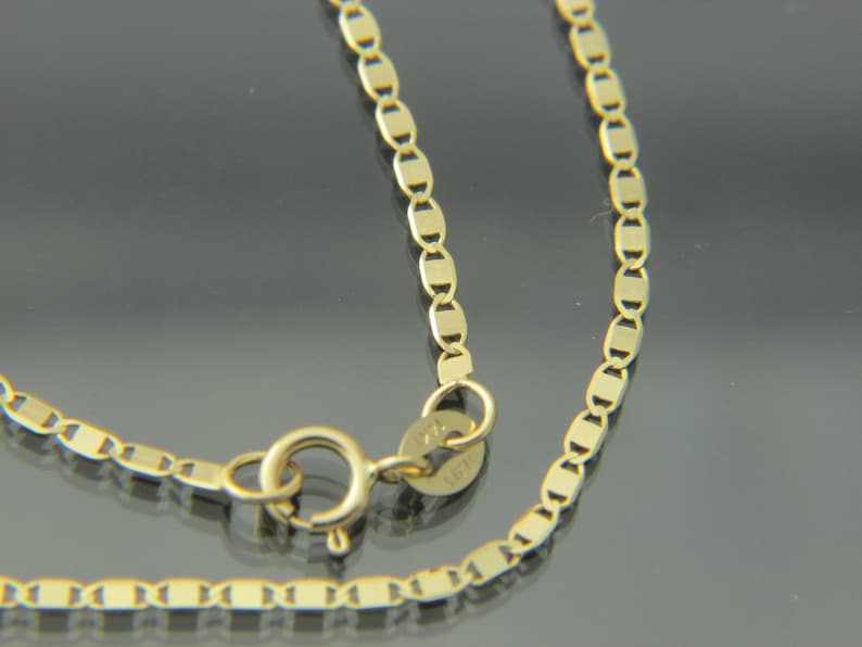 May include: A close-up of a gold necklace with a delicate chain design. The chain features small, rectangular links connected in a repeating pattern. The necklace has a spring ring clasp for secure closure.